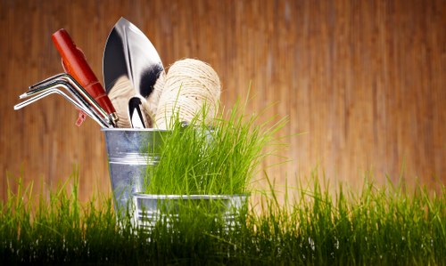 An outdoor scene featuring a collection of gardening tools and supplies placed in a metal bucket, including a trowel, a hand rake, a wooden-handled gardening fork, and a spool of natural twine, with a bundle of fresh green grass or plants nearby. The background consists of a warm, wooden surface, suggesting a garden shed or outdoor workspace. The foreground shows a lush, vibrant green lawn with evenly cut grass, typical of well-maintained garden areas in Ilford, with natural daylight highlighting the textures of the grass, tools, and wooden backdrop. This image emphasizes basic gardening equipment and a tidy outdoor environment, which aligns with professional gardening and lawn care services provided by Lawn Mowing Ilford and other local providers in the area, supporting the relevance to outdoor maintenance, turf care, and garden upkeep services in the postcodes around Ilford.