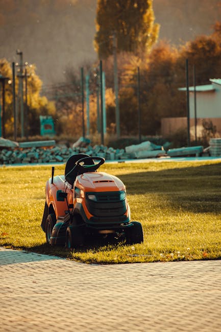 A bright orange lawnmower with a black steering wheel and protective cover is positioned on a neatly trimmed grassy area of a large garden. The lawnmower appears to be a ride-on type, suitable for ext